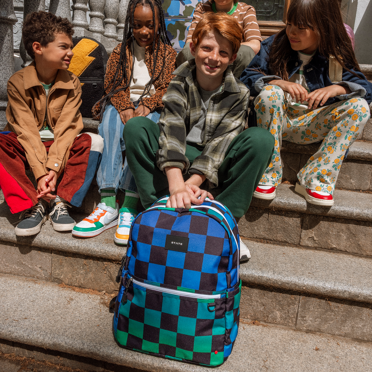 boy sitting on stairs holding Kane double pocket backpack blue checkerboard